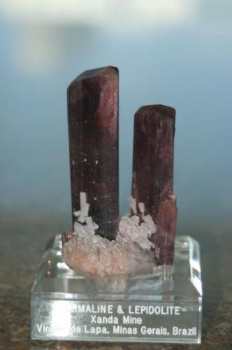 One of my favorites. 2 dark red / cranberry tourmalines on lepidolite matrix.  Both of these have gemmy tops, and perfect terminations. The smaller xtal has one repair. This piece is from Xanda Mine, Virgem de Lapa, Minas Gerias Brazil. Dimensions are 8.9 x 5.1 x 5 cm and weight is 105 grams. One picture is in the case with backlight. (Author: VRigatti)