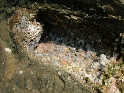 Thomsonite + Apophyllite
Moonen Bay, Isle of Skye, Scotland, UK
FOV approx 50cm across
A large pipe cavity lined with apophyllite and stalactitic thomsonite. The pipe is about 20cm in diameter. An unusual occurence; haven’t seen another one like it at Moonen Bay or elsewhere. Other cavities in the same boulder contain the same minerals, but in completely different habits to this single example.
Photo taken April 2005. (Author: Mike Wood)