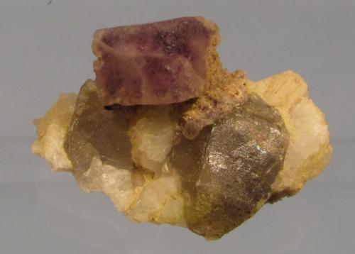 Fluorite on Albite and Smoky Quartz
Lundy Island, Devon, England, UK
Specimen 24mm x 14mm tall
A similar specimen to the previous, the purple fluorite ’cube’ is slightly smaller, also slightly etched and with corner modifications. There is a tiny (1mm) topaz crystal just below on the right of the fluorite crystal; interestingly under the microscope it appears to have a purple fluorite crystal included within! (Author: Mike Wood)