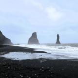 Al este de los acantilados de Reynisfjall, se encuentra la playa de ‘arena negra’ de Reynisfjara, en Vík í Mýrdal. La playa está formada por pequeños fragmentos negros y brillantes de basalto, de unos 2-3 mm, como producto del rápido enfriamiento del magma en contacto con el agua, que hace que se descomponga en trozos más pequeños, y de la fuerte erosión marina posterior. Además de basalto, la arena negra está formada por restos de piroxenos (augita), olivino, plagioclasa (labradorita), magnetita e ilmenita, en cantidades variables.
En la imagen, dos agujas de basalto en la playa de Reynisfjara, conocidas como Reynisdrangar. Miden 43 metros de altura y son el resultado de la erosión del material más blando que rodeaba al basalto más duro. Para los que vieron la serie de HBO &rsquo;Juego de Tronos&rsquo;, Reynisdrangar aparece en un capítulo de la séptima temporada. (Autor: Antonio P. López)