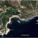AERIAL PHOTOGRAPH – TERRANERA MINE SITE

This aerial view captures a vibrant tableau of East Elba&rsquo;s coastline, a place where the echoes of a laborious industrial past now serve as a dramatic backdrop to modern leisure. The landscape is dominated by two key tourist landmarks: the shimmering, dark waters of Laghetto di Terranera, a small coastal lake, and the adjacent Spiaggia di Terranera, its shoreline a lively mosaic of colourful umbrellas and the joyful activity of sunbathers enjoying the Tyrrhenian Sea.

Beneath this idyllic scene, a deeper history is etched into the very rock and soil. At the location marked “A,” the skeletal remains of a structure from the island&rsquo;s mining era stand as a silent testament to a bygone age. The dilapidated steel frame and decaying brick walls, spectral remnants of a bygone industry, loom over the water. Below them, a solitary stone foundation, a testament to the structure&rsquo;s original footprint, juts into the sea. Its stones, once a firm base, are now worn smooth by the ceaseless caress of the waves, a quiet sentinel marking the inexorable passage of time. Further inland, the subtle depression highlighted at “B” marks a portion of the former Capo Bianco mine. This great wound in the earth has long since been filled and secured, a scar healed over by time and reclamation efforts, leaving only a gentle concavity in the terrain to hint at the excavation that once took place.

The geological narrative of this coastline is written in the dramatic contrast of its rock formations. The foundational rock is dark, layered Schist, which provides a somber canvas for the intrusions that give the area its mineral wealth. Cutting through this ancient metamorphic rock are dykes of upper Miocene leucogranites —intrusive veins of pale, crystalline igneous rock that forced their way up from the Earth&rsquo;s mantle. These bright granitic fingers are prominent from the area just north of Capo Bianco, sweeping northwards, and re-emerging dramatically as a major outcrop that breaks the sea&rsquo;s surface approximately 100 metres from the Cape itself. Adjacent to these outcrops are lenses of mylonite marble.

This very geological intrusion is the source of the region&rsquo;s famed Iron. The mineralization presents itself in two distinct patterns. At the site of the Capo Bianco mine, the valuable ores were concentrated in a dense, egg-shaped pod, a geological prize that was vigorously exploited. A more extensive and visually striking formation traces a vast, horse-shoe shaped halo around the Laghetto di Terranera. This rust-coloured band of mineralization, approximately 50 metres in width, follows the western and eastern shorelines of the lake and beyond, a tell-tale stain revealing the rich deposits of iron that once made this stunning coastline a centre of intense industrial labour. (Author: silvia)